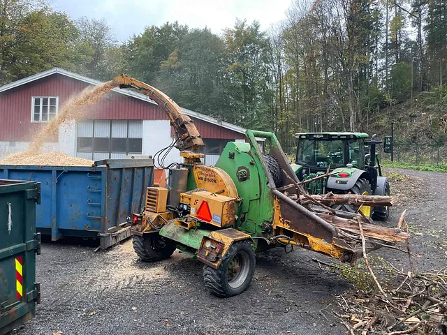 Flishugger mater greiner inn og blåser flis i blå container foran rød bygning.