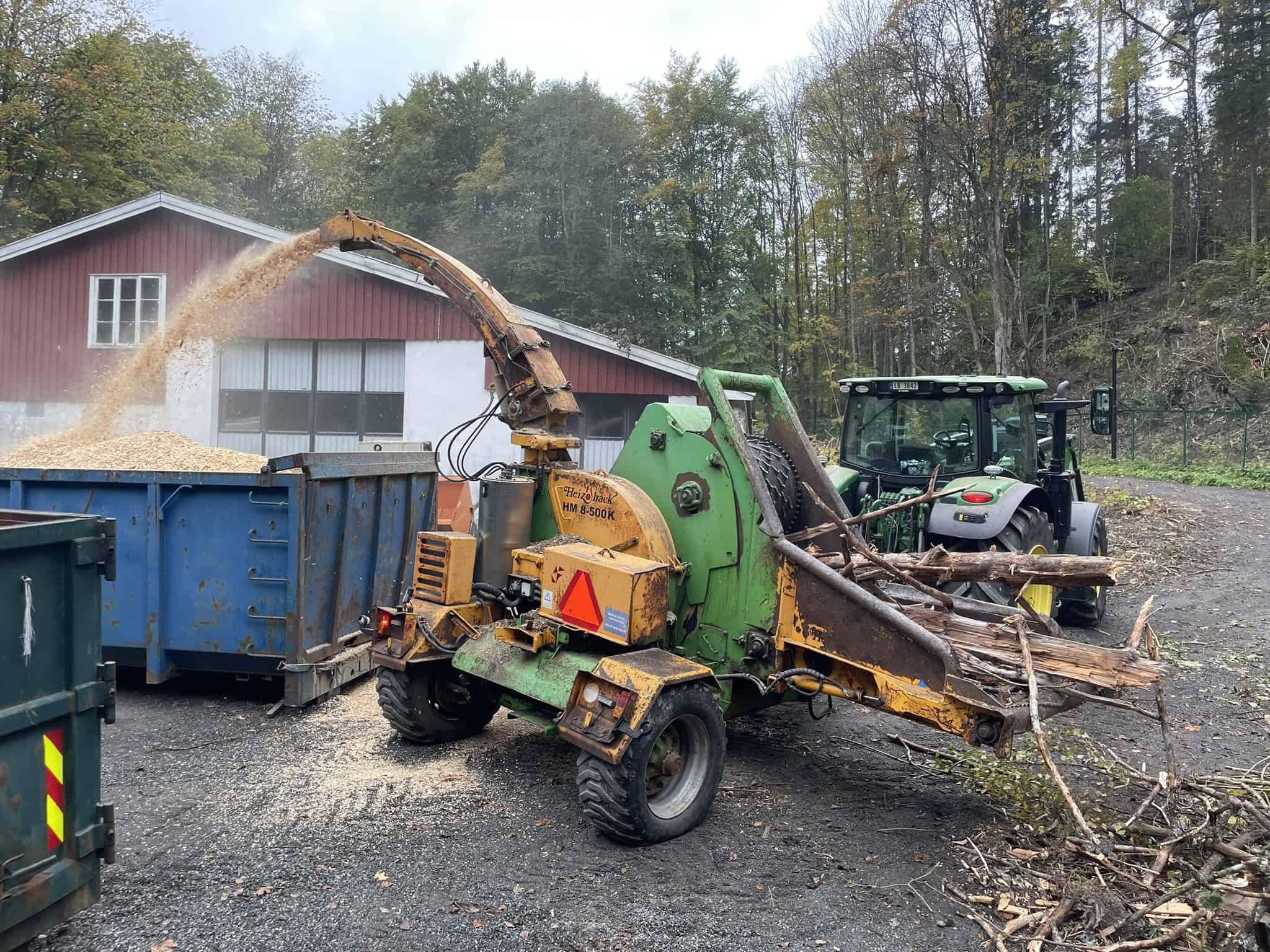 Flishugger mater greiner inn og blåser flis i blå container foran rød bygning.