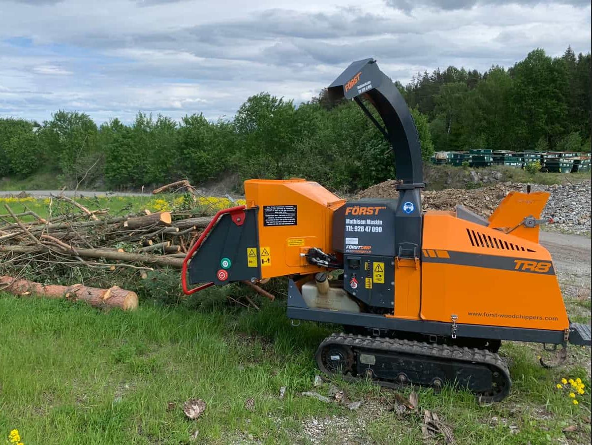 Flismaskin med belter maler opp kvister og stokker ved en grønn veiskråning nær skog og grustak.