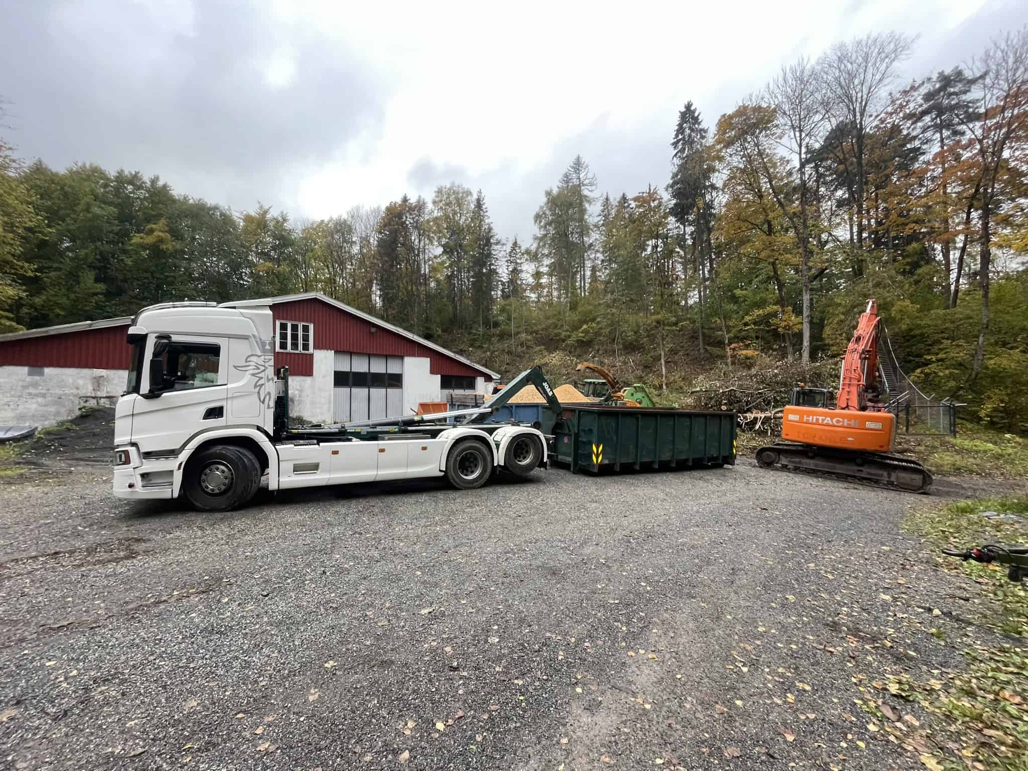 Hvit lastebil og oransje gravemaskin foran rød bygning med trefelling i bakgrunnen.