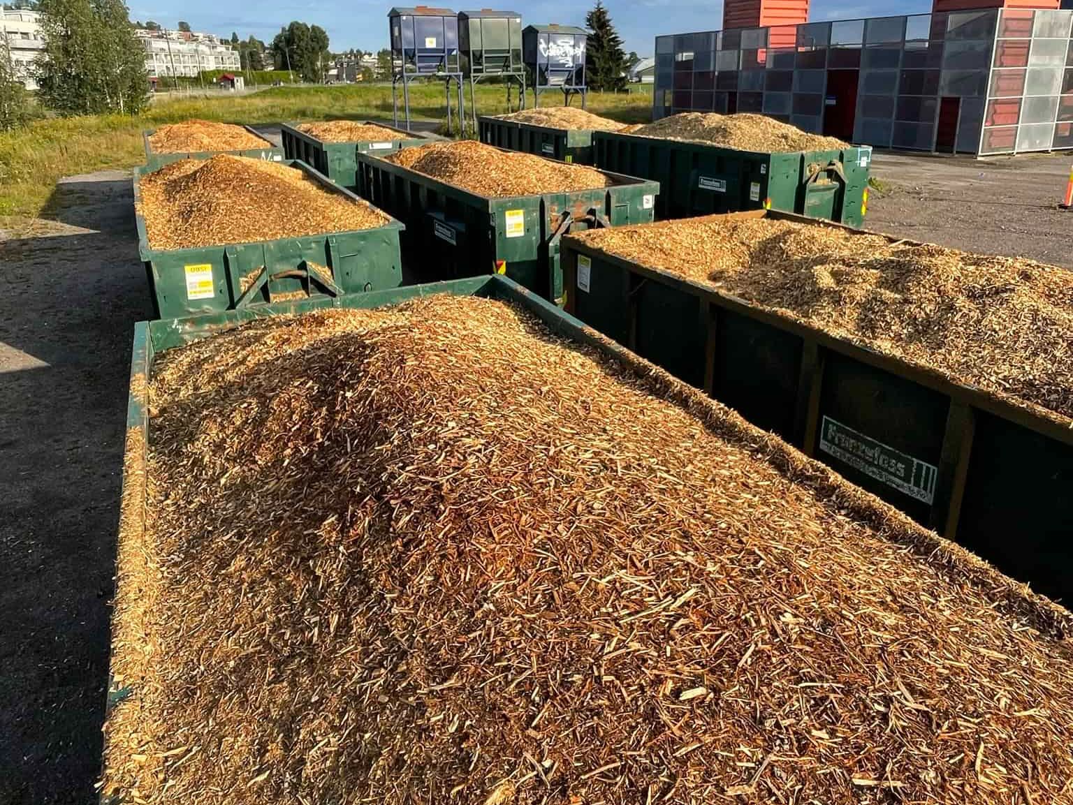 Store containere fylt med flis står utendørs ved industriområde under blå himmel.
